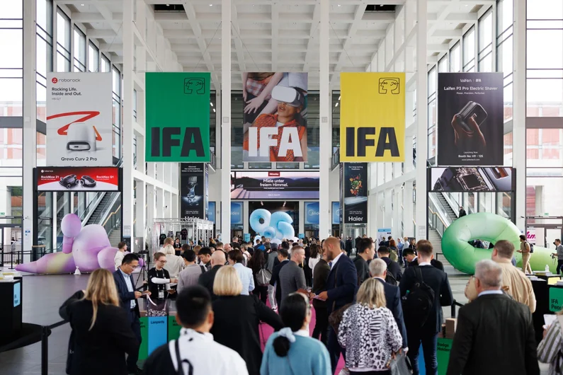 A crowded indoor venue at the IFA Berlin technology expo, featuring colorful banners, product displays, and attendees walking around.