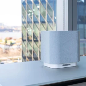 A sleek smart speaker with a light gray fabric finish on a desk near a large office window. Glass building and urban landscape in the background.