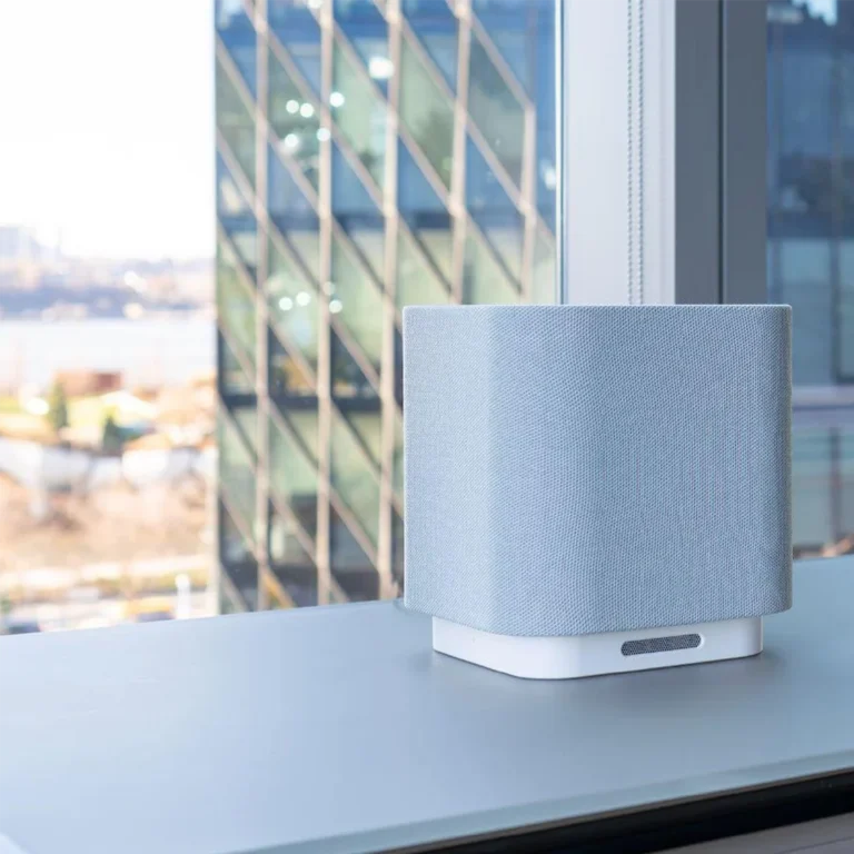 A sleek smart speaker with a light gray fabric finish on a desk near a large office window. Glass building and urban landscape in the background.