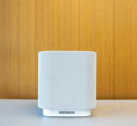 A cylindrical smart speaker in light gray fabric with minimalist white base, placed on a table with a wooden backdrop.