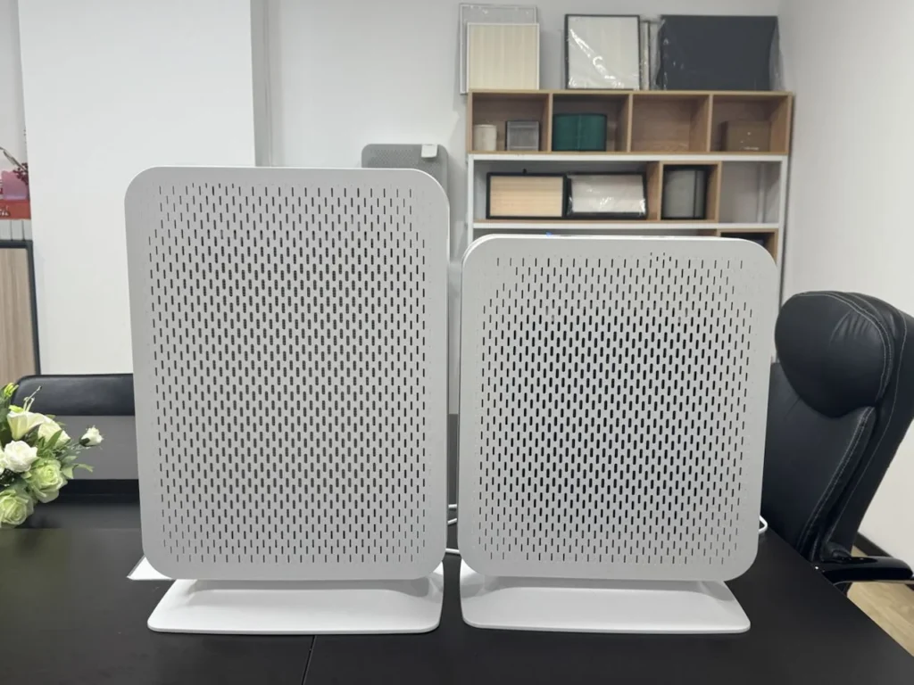 Two white air purifiers side by side on a black table in an office with shelving and a chair in the background.