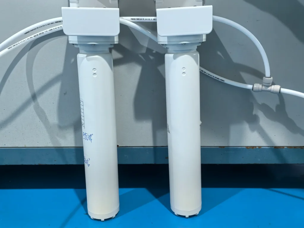 Close-up of a water filtration system with two cylindrical white filter housings connected to white pipes against a blue and gray background.