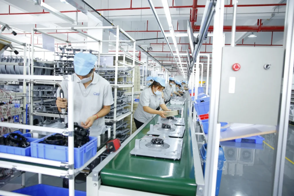 A group of workers in uniforms and caps assembling products on a factory assembly line with blue bins and industrial equipment.